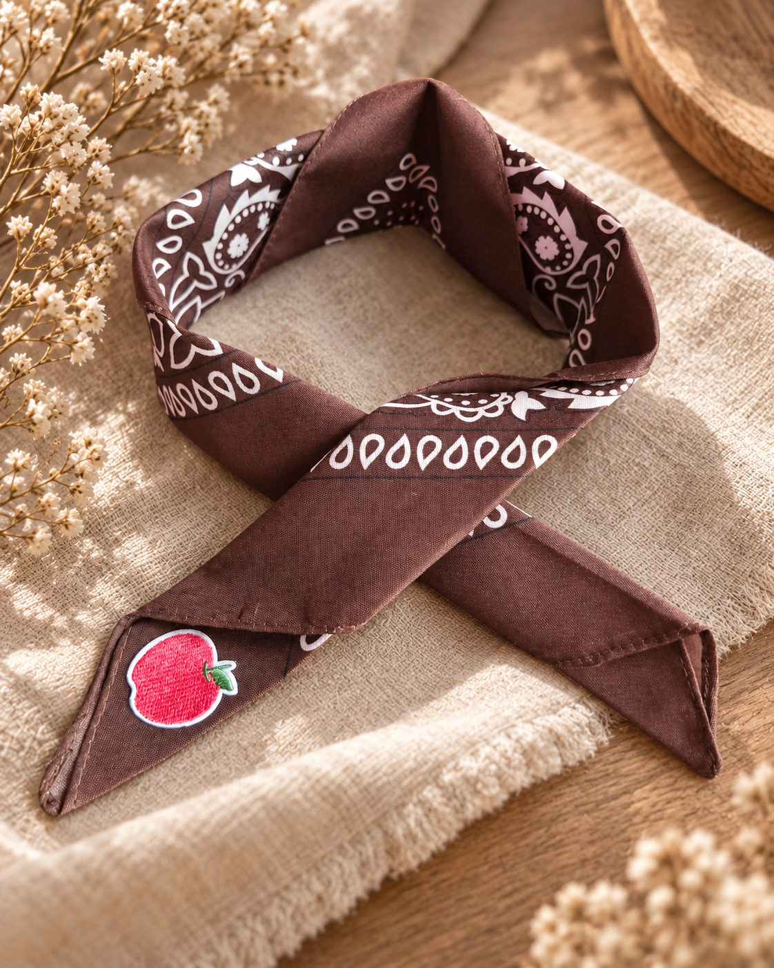 Apple Brown kinderbandana in chocoladebruine paisley, opgevouwen met een embleem van een rode appel op de punt